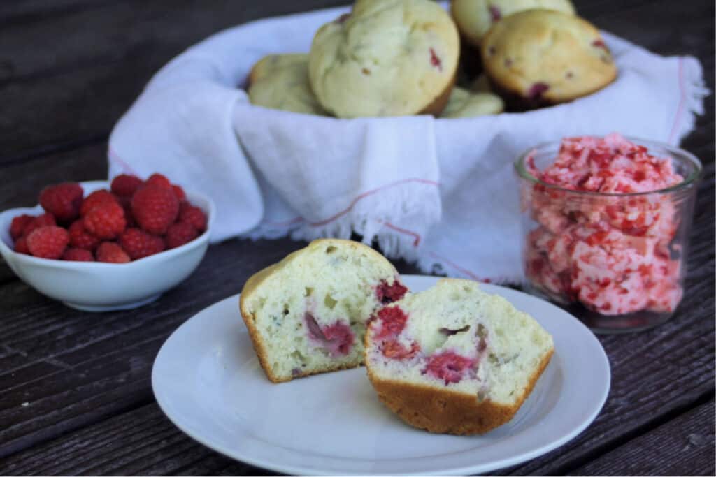 A raspberry cream cheese muffin sliced in half on a white plate sitting in front of a basket full of muffins and raspberry butter.