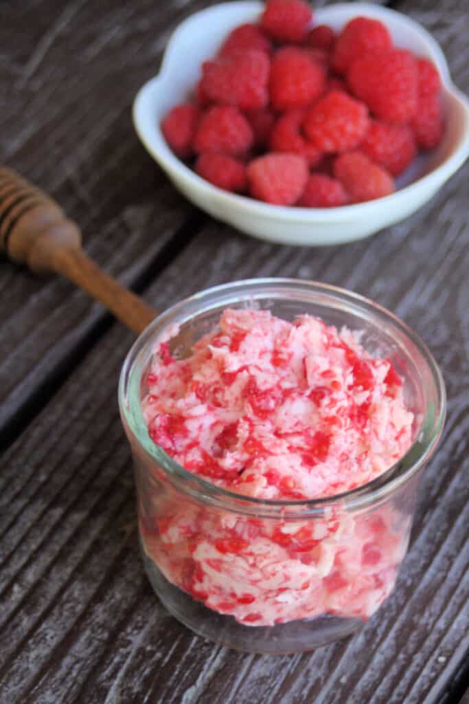 An open jar of raspberry honey butter sitting in front of a white bowl full of fresh raspberries