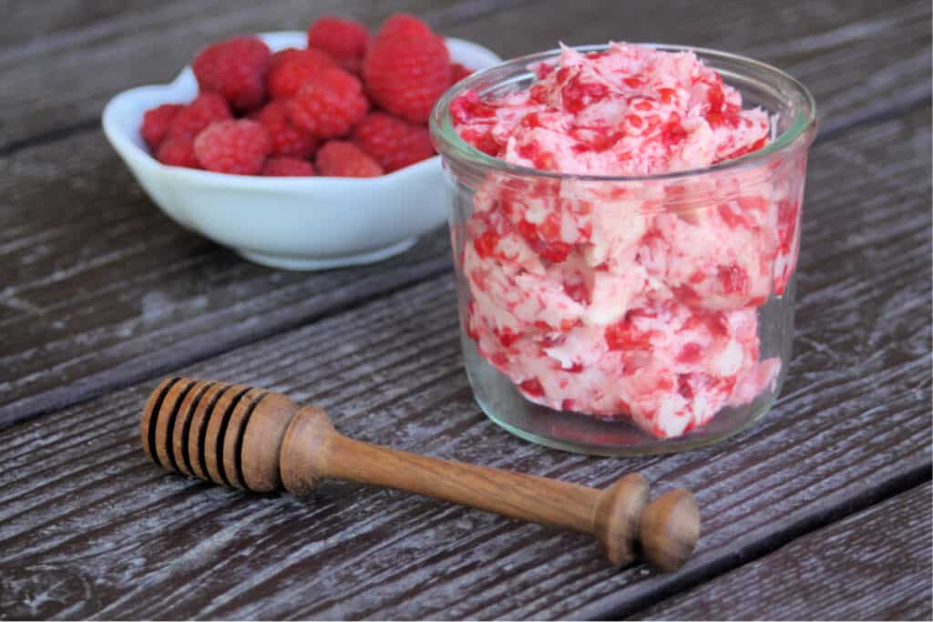 Raspberry honey butter in an open glass jar sitting behind a honey dipper and in front of  a white bowl full of fresh raspberries. 