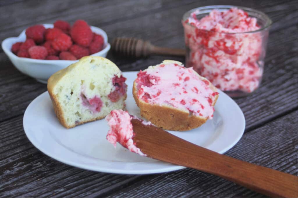 A raspberry muffin sliced in half on a white plate - one half is spread with raspberry honey butter. A wooden spreader sitting on the plate. 