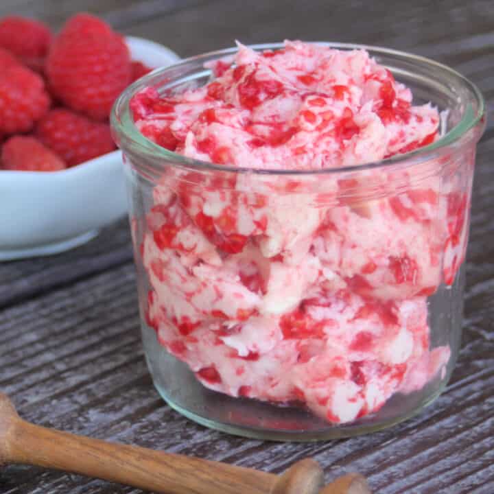 An open jar full of raspberry honey butter sitting in front of a white bowl full of fresh raspberries.