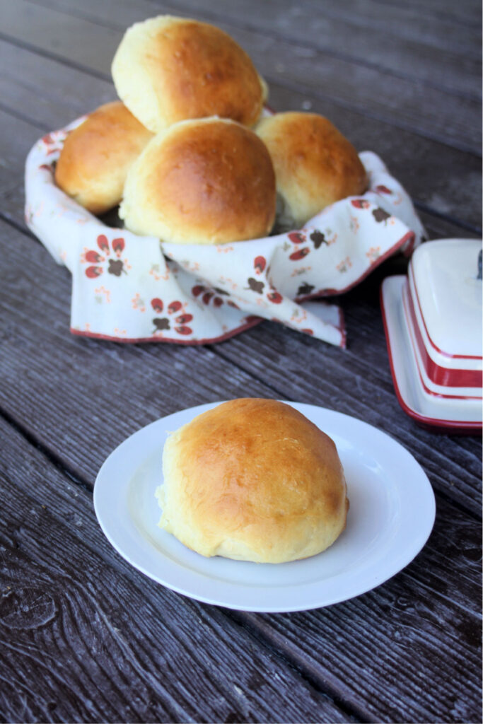 A sweet potato bun on a white plate sitting in front of a basket full of more buns and a butter dish.