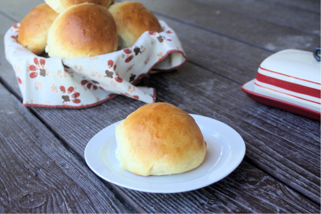 A sweet potato bun on a white plate sitting in front of a basket full of more buns and a butter dish.