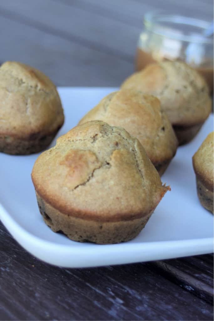 Apple butter muffins on a white plate sitting in front of an open jar of apple butter with a spoon sitting inside