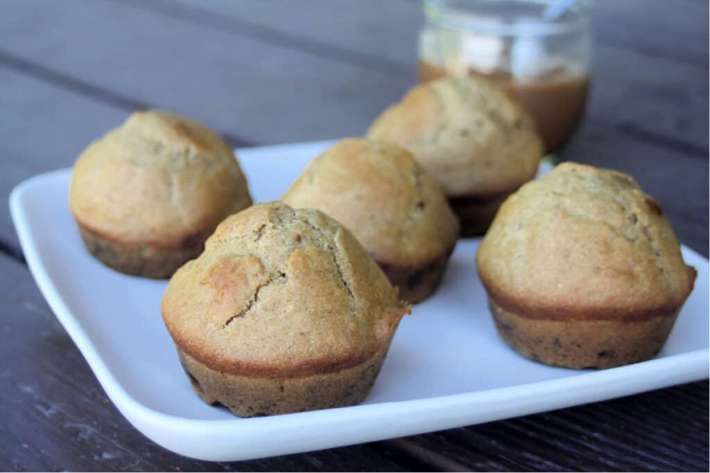 Apple butter muffins on a white plate sitting in front of an open jar of apple butter with a spoon sitting inside