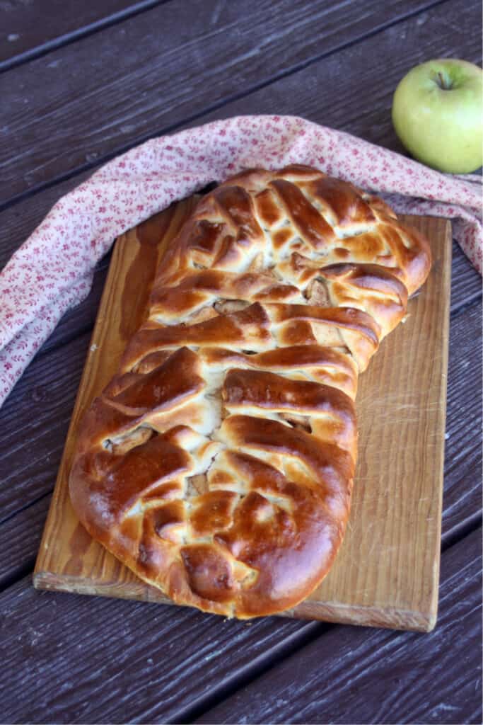 An apple cream cheese braid on a wooden cutting board with a napkin behind it and a green apple.