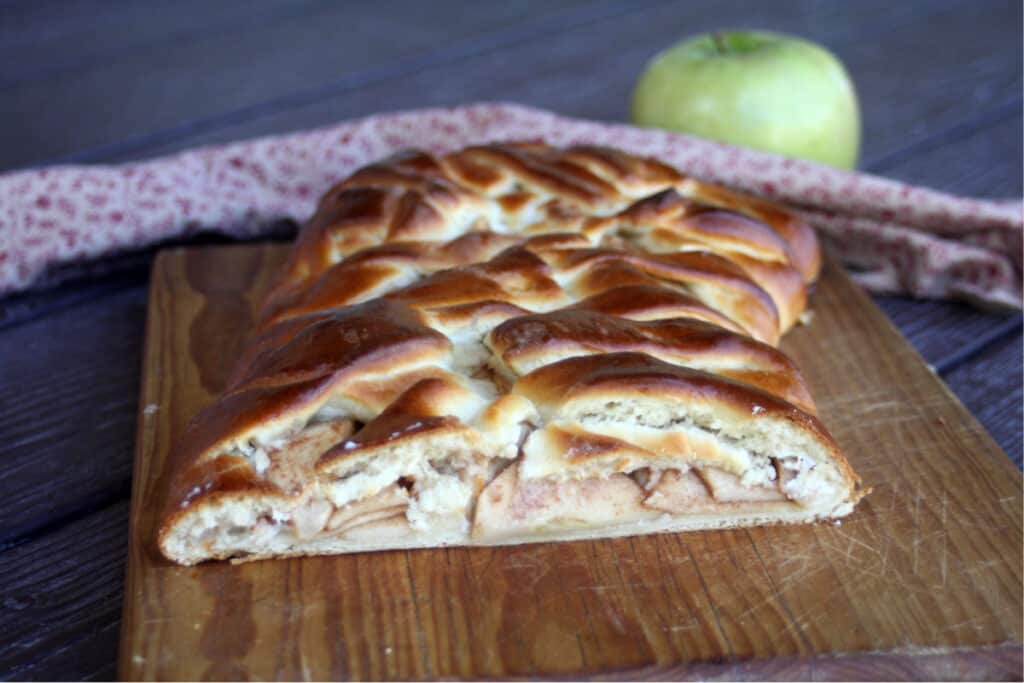 An apple cream cheese braid with the end cut off exposing the inside sitting on a wooden cutting board with a napkin and green apple sitting in the background. 