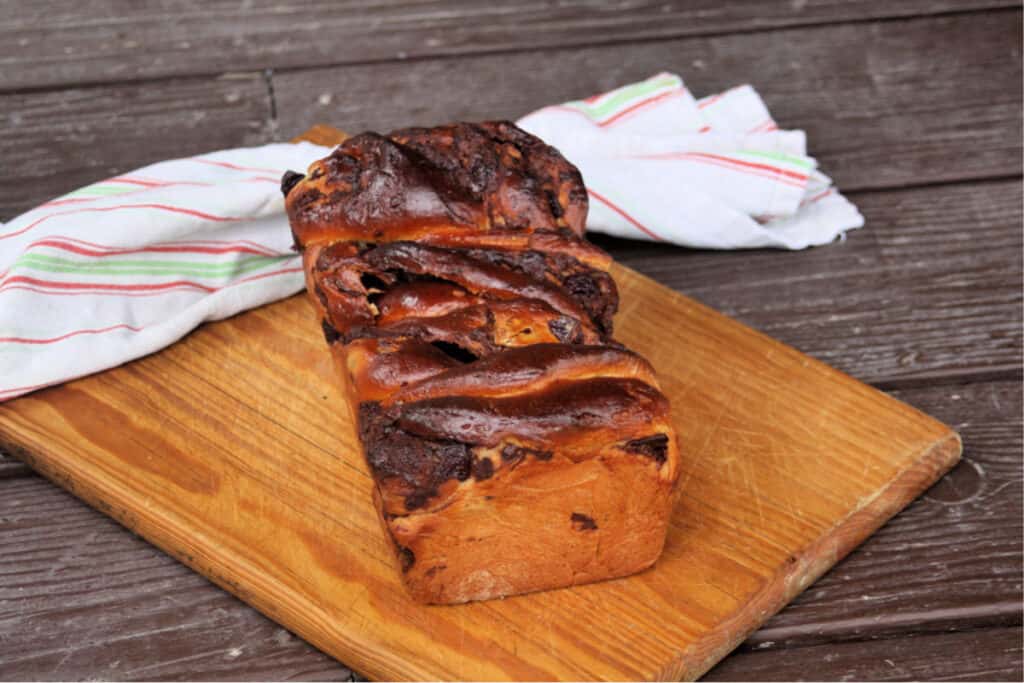 A loaf of chocolate cherry babka on a wooden cutting board with a towel draped across the back of the board.