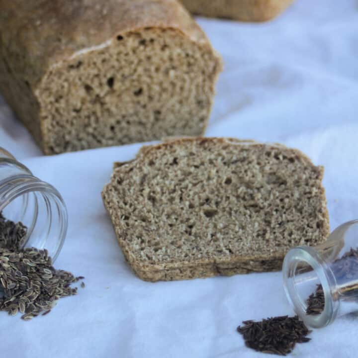 A slice of dill rye bread on a white cloth surrounded by seeds and loaf of bread in the background.
