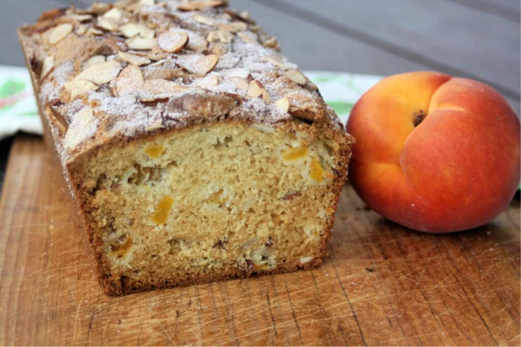 A loaf of peach bread with the end cut off exposing the inside on a cutting board sitting next to a fresh peach.