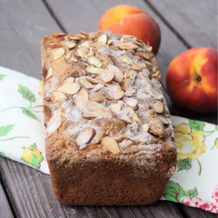 A loaf of peach bread sitting on a floral table runner in front of two fresh peaches.