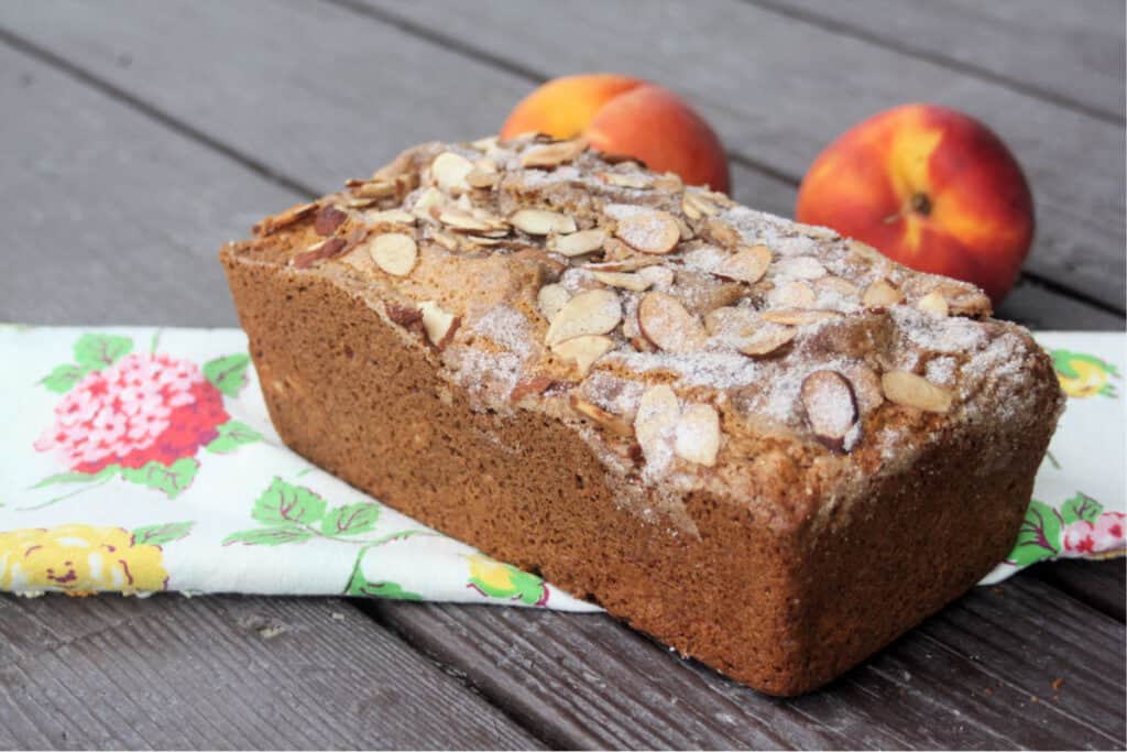 A loaf of peach bread as seen from the side sitting on top of a floral table runner in front of 2 fresh peaches. 