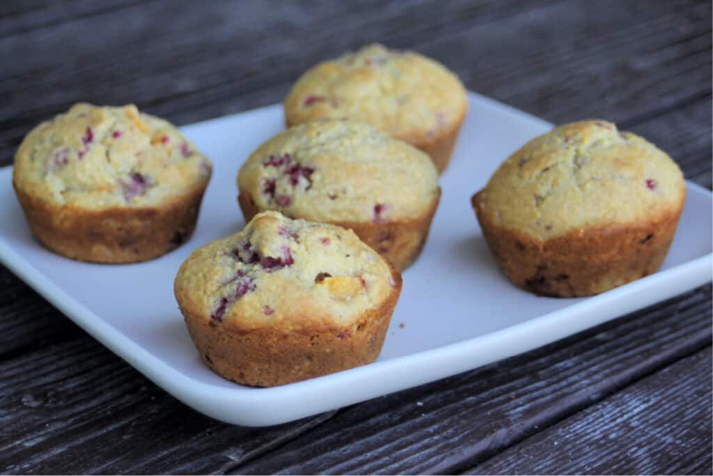 A white plate with five peach raspberry cornmeal muffins spread on it.