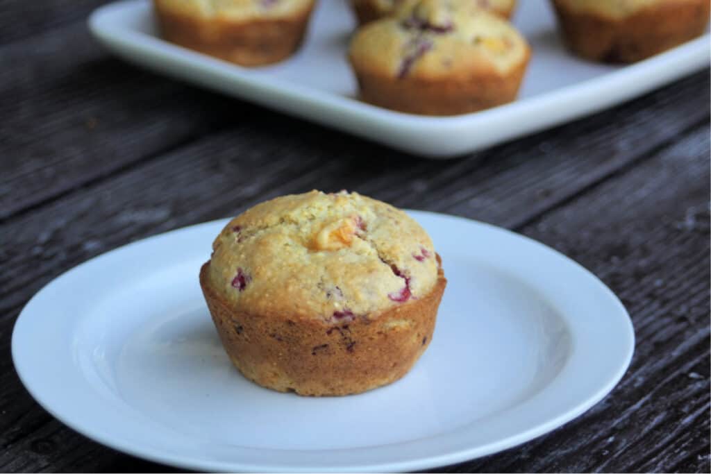 A peach raspberry muffin on a white plate sitting in front of a platter full of muffins.