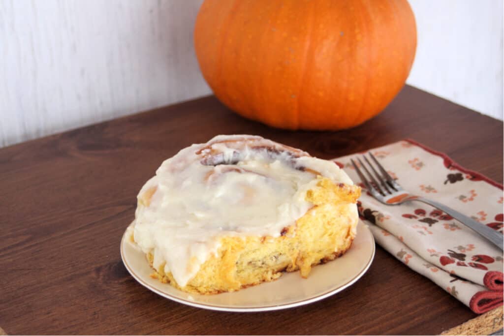 A pumpkin cinnamon roll with cream cheese frosting on a white plate sitting next to a napkin with a fork on top. A whole pumpkin sitting behind. 