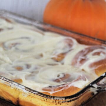 A glass casserole dish full of Pumpkin cinnamon rolls with cream cheese frosting sitting on a wooden cutting board in front of a pumpkin