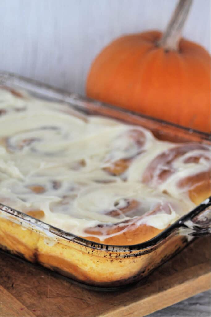 A glass casserole dish full of Pumpkin cinnamon rolls with cream cheese frosting sitting on a wooden cutting board in front of a pumpkin.