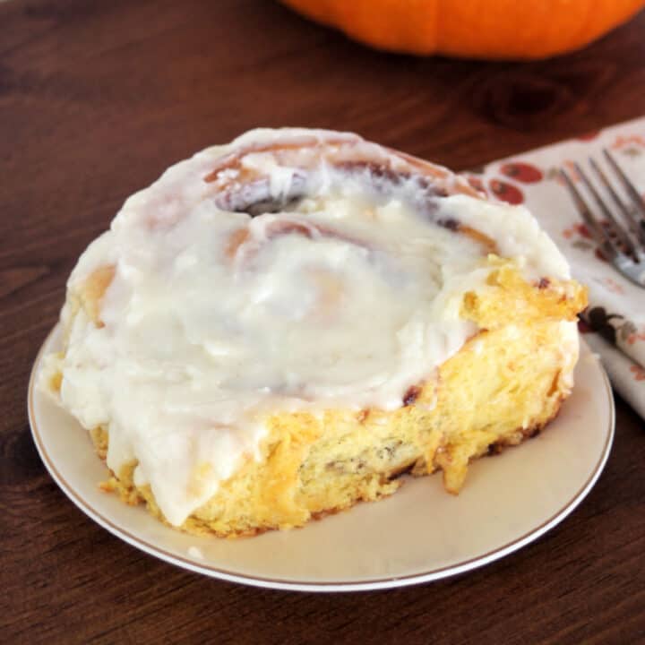 A pumpkin cinnamon roll with cream cheese frosting on a white plate sitting next to a napkin with fork on top.