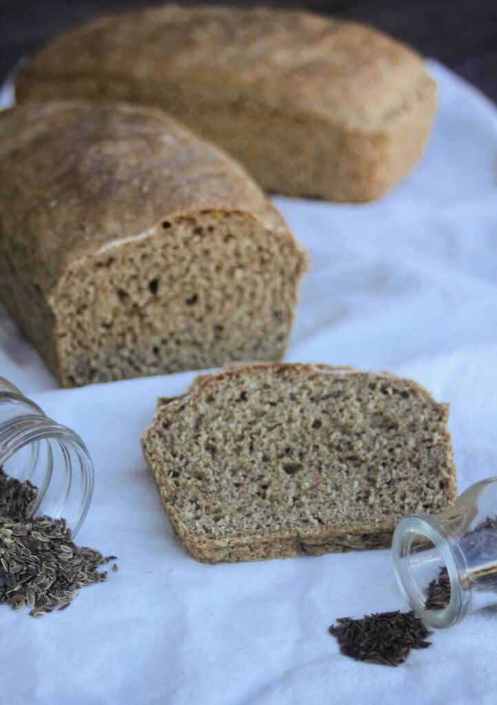 A slice of dill rye bread sitting behind jars of seeds on a white cloth, rest of loaf in the background. 