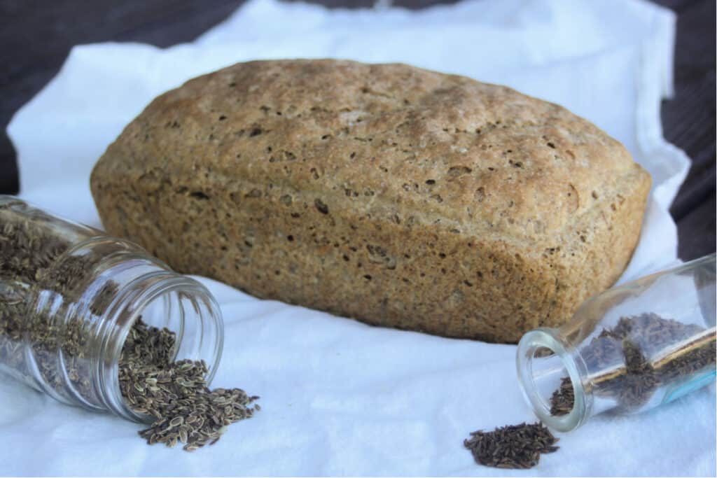 A loaf of dill rye bread sitting on a white cloth with jars of seeds in front. 