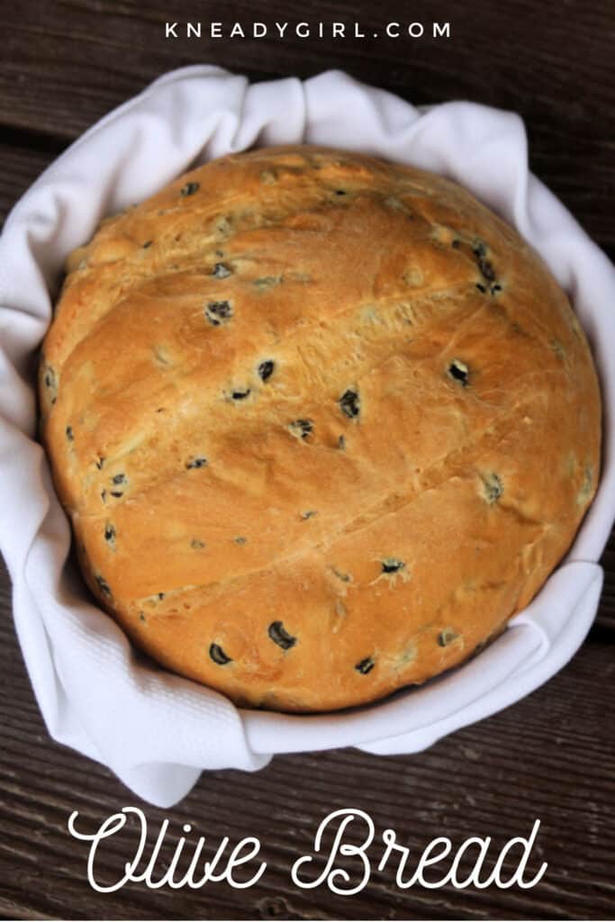 A round loaf of olive bread in a white linen lined basket with the words olive bread on the bottom.