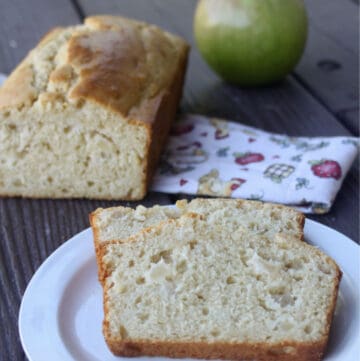 slices of apple bread on a white plate sitting front of the rest of the loaf and a fresh green apple.