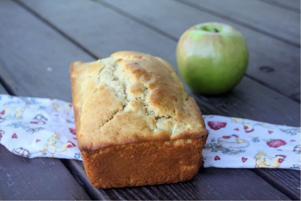 A loaf of apple bread sitting on a linen with green apple in the background.