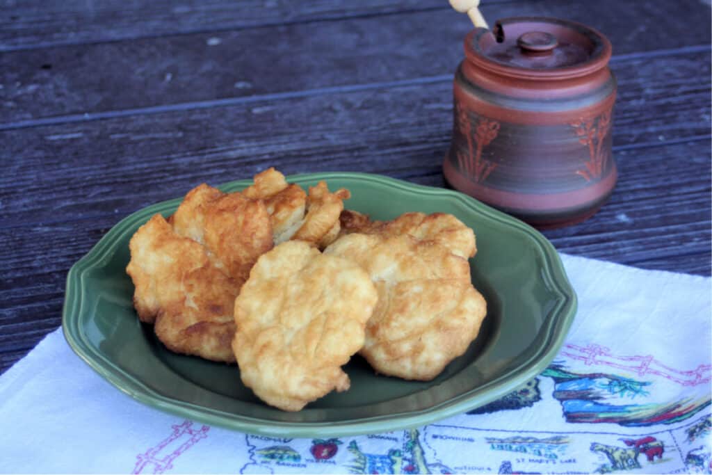 Pieces of fry bread stacked on a green plate sitting on a white linen with a clay honey pot sitting behind them.