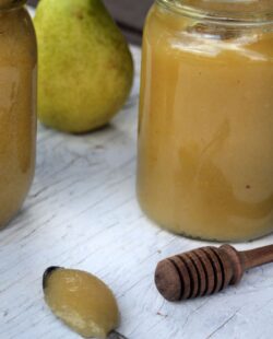 Pear butter on a spoon sitting in front of jars of more pear butter, a honey dipper and fresh pears.