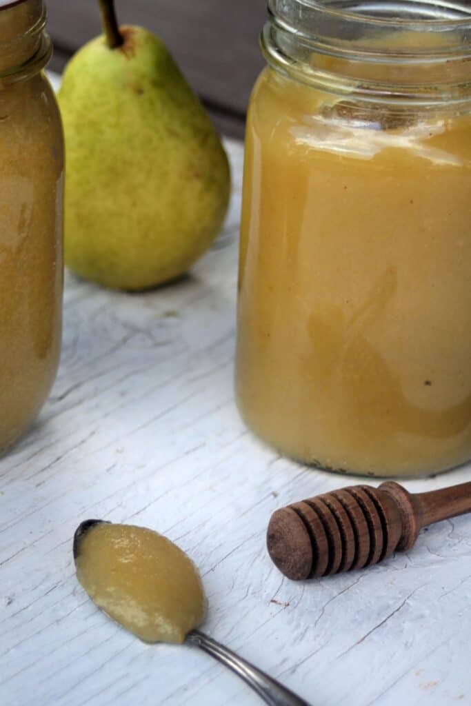 Pear butter on a spoon sitting in front of jars of more pear butter, a honey dipper and fresh pears.