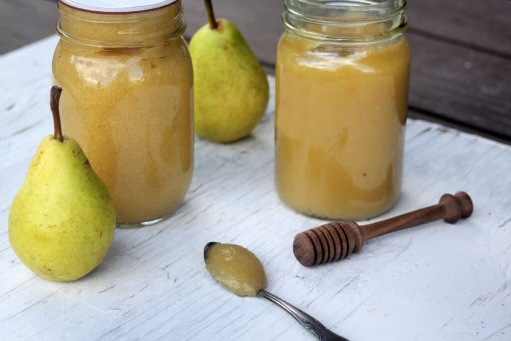 Pear butter on a spoon sitting in front of jars of more pear butter, a honey dipper and fresh pears.