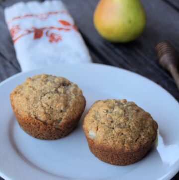 2 muffins on a white plate with a napkin, fresh pear, and honey dipper in the background.