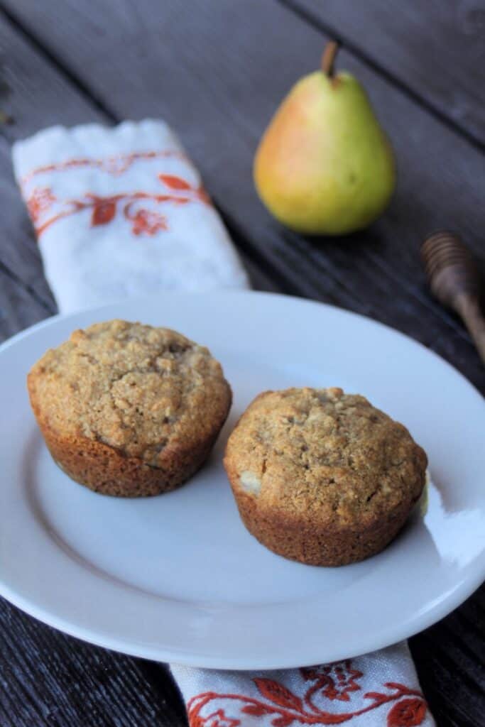 2 muffins on a white plate with a napkin, fresh pear, and honey dipper in the background.