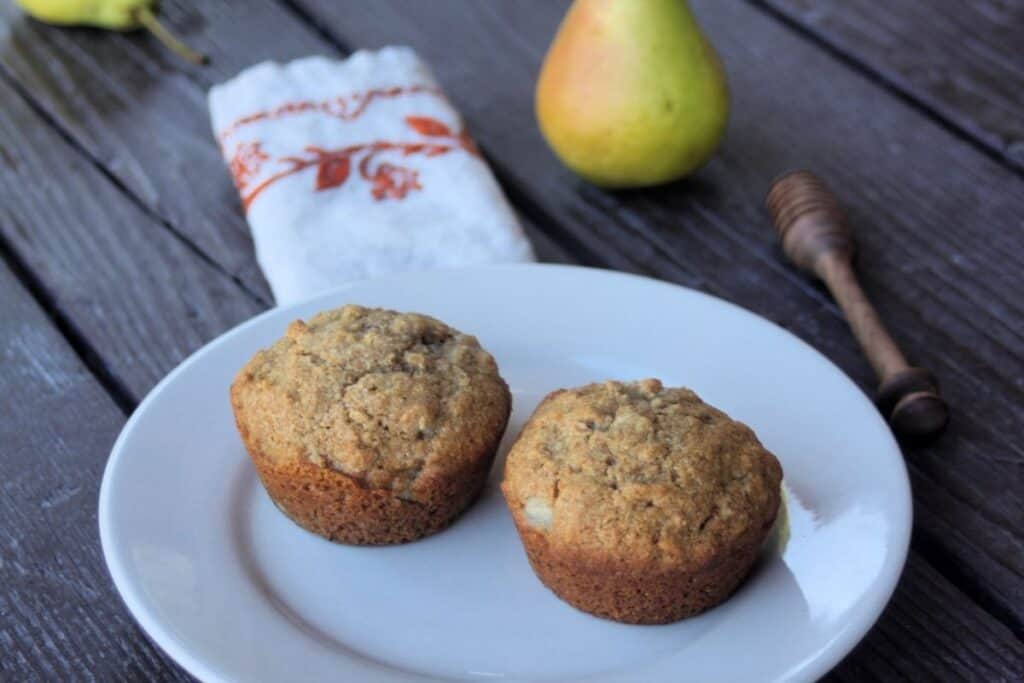 2 muffins on a white plate with a napkin, fresh pear, and honey dipper in the background.