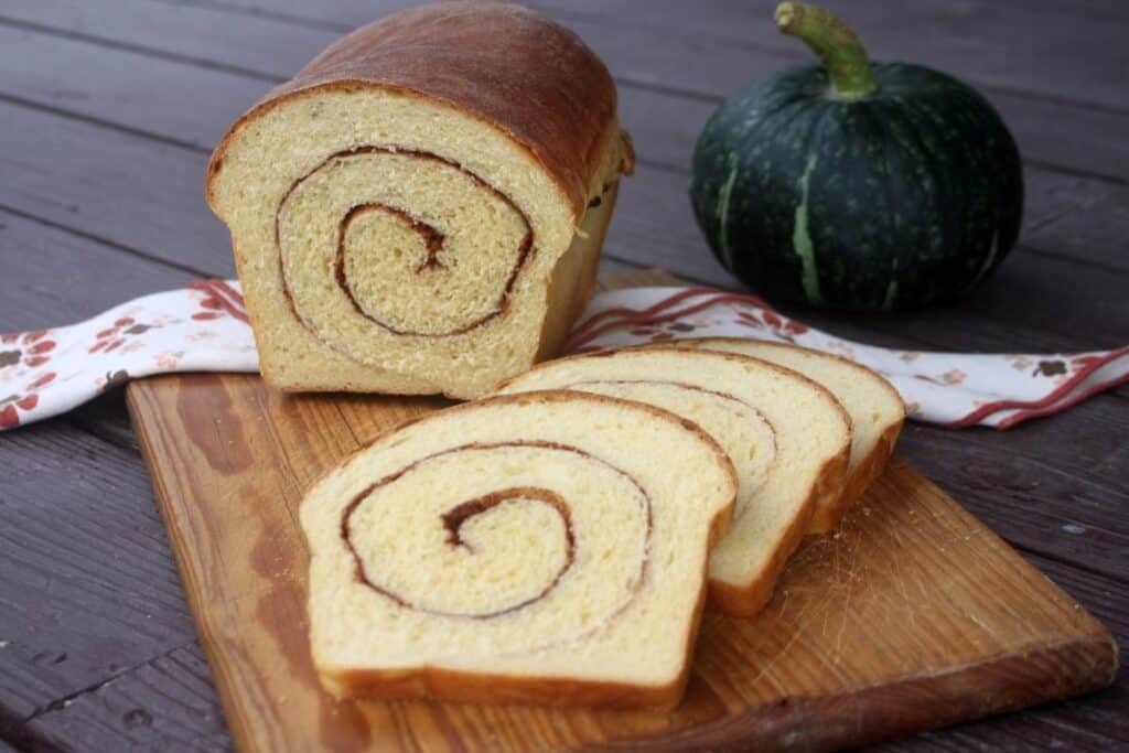 Slices of pumpkin cinnamon swirl bread on a cutting board with remaining loaf behind them and a green pumpkin in the background.