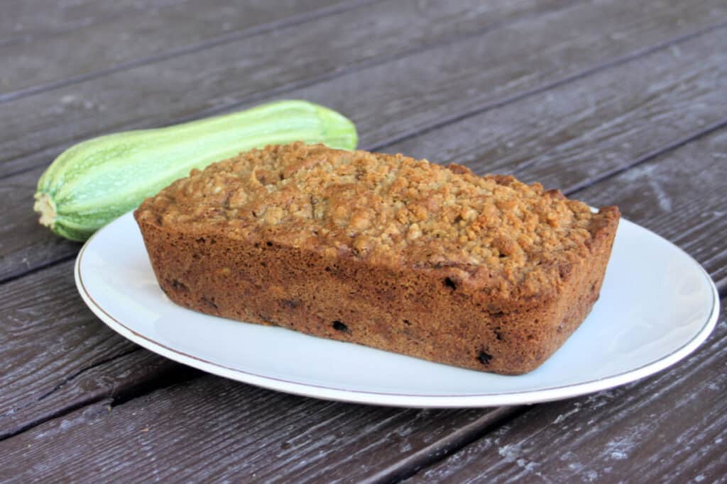 An entire loaf of zucchini raisin bread on a white platter with a whole fresh zucchini sitting to the left.