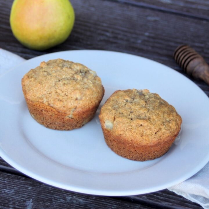 2 muffins on a white plate with a napkin, fresh pear, and honey dipper in the background.