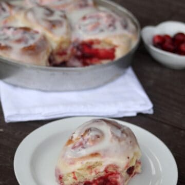 A cranberry sweet roll sitting on a white plate with a napkin sitting underneath a pan full of rolls and a white dish full of fresh cranberries in the background.