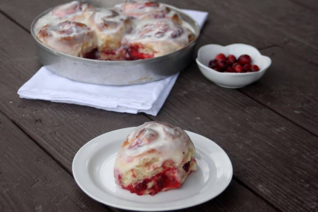 A cranberry sweet roll sitting on a white plate with a napkin sitting underneath a pan full of rolls and a white dish full of fresh cranberries in the background.