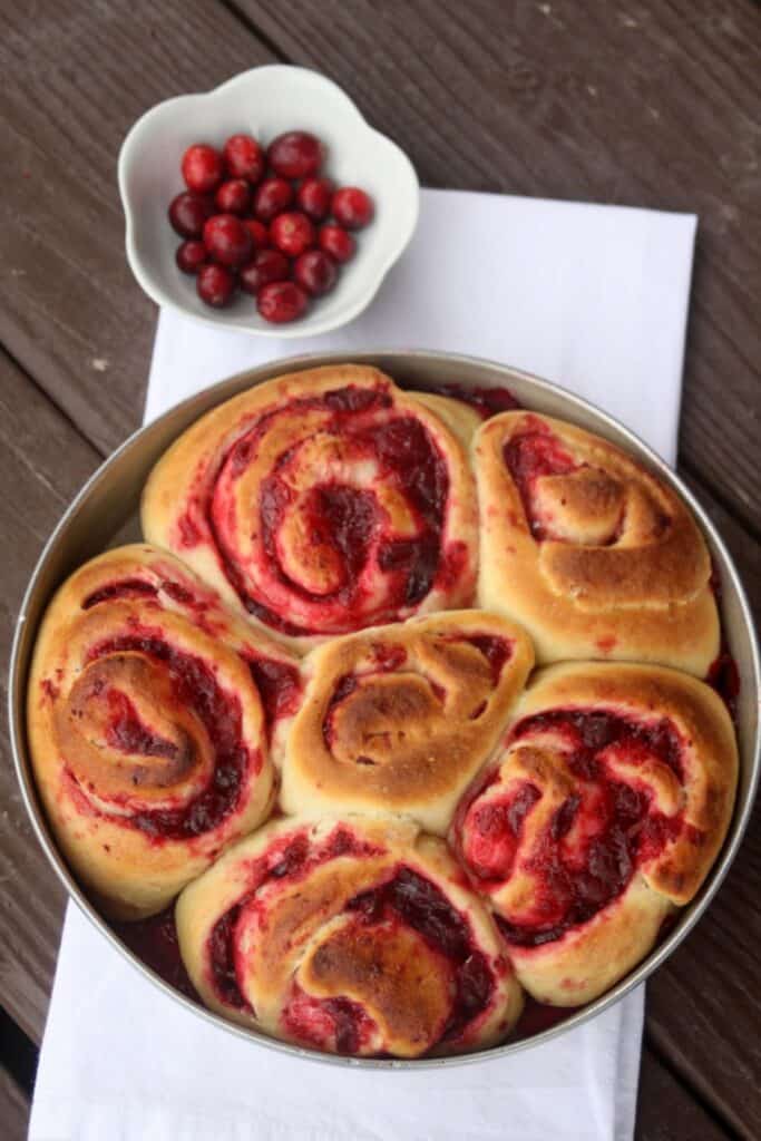 A pan of unfrosted cranberry sweet rolls sitting on a white towel with a bowl full of fresh cranberries.
