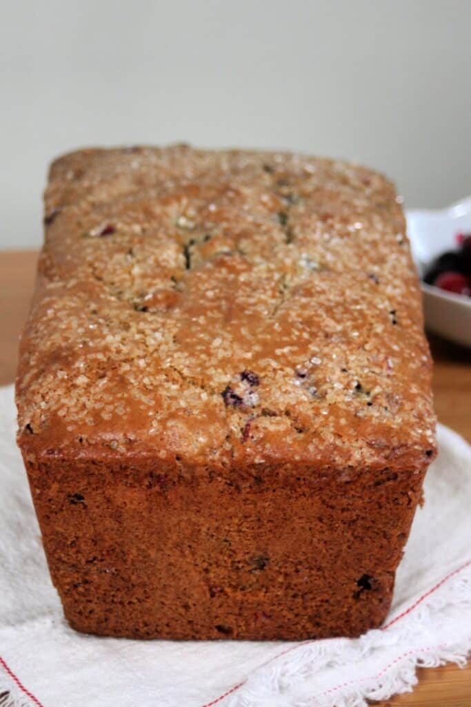 A slice of cranberry bread on a white plate sitting in front of the remaining loaf and a bowl of cranberries.