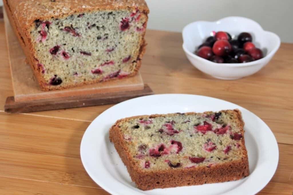 A slice of cranberry bread on a white plate sitting in front of the remaining loaf and a bowl of cranberries.