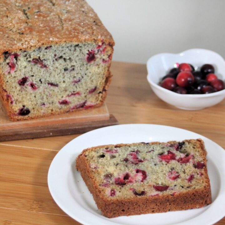 A slice of cranberry bread on a white plate sitting in front of the remaining loaf and a bowl of cranberries.