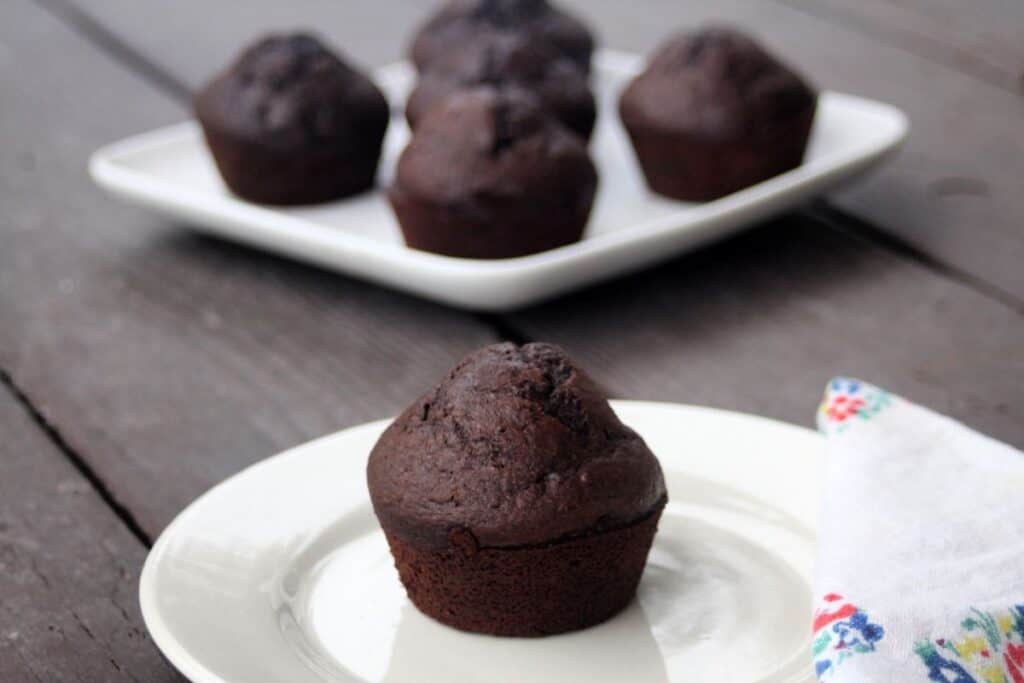 A malted chocolate muffin sitting on a plate with a floral napkin next to it and a platter of muffins behind. 