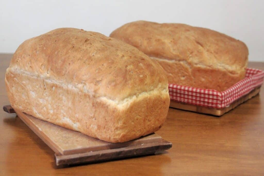 A loaf of cream bread sitting on a cutting board with another sitting behind it in a red and white checked lined basket.