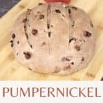 A round loaf of bread on a wood cutting board with a red heart shaped container sitting behind it and text overlay reading: pumpernickel raisin bread.