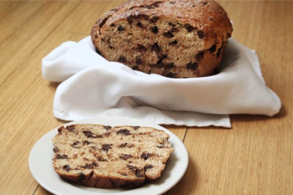 A slice of chocolate chip bread on a plate with remaining loaf sitting in a linen lined basket in the background.