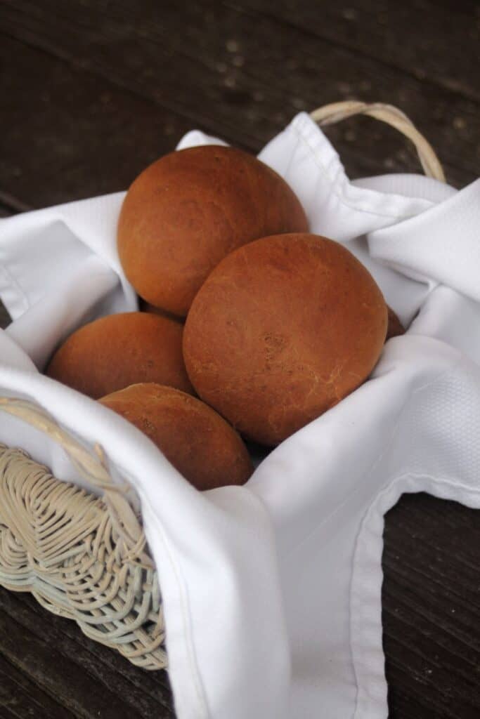 Molasses brown bread rolls stacked in a white napkin lined basket.