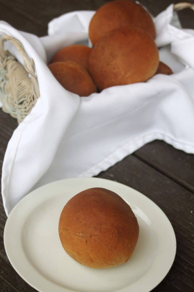 a molasses brown bread dinner roll on a white plate sitting in front of a napkin lined basket full of more rolls.