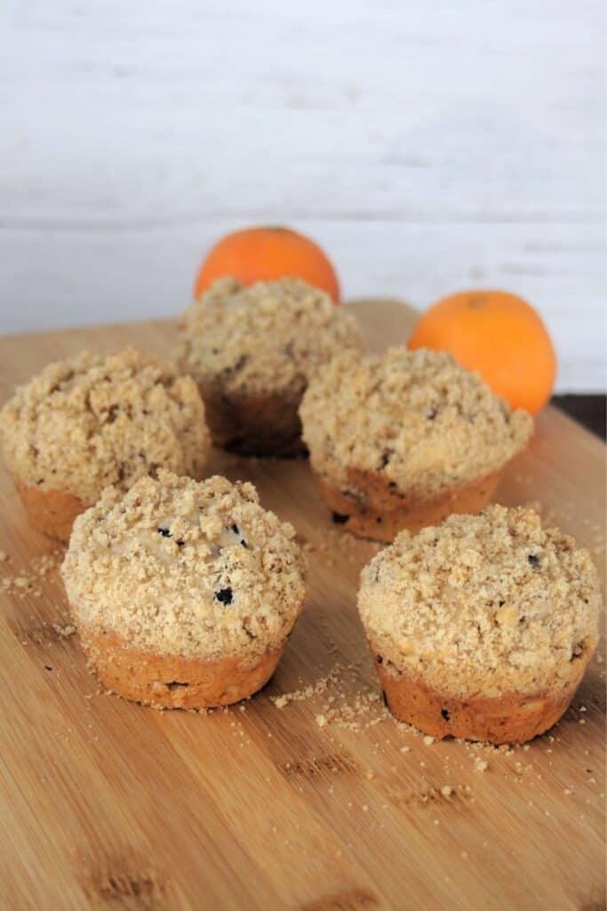 Dried cranberry muffins lined up on a wooden cutting board with oranges in the background.